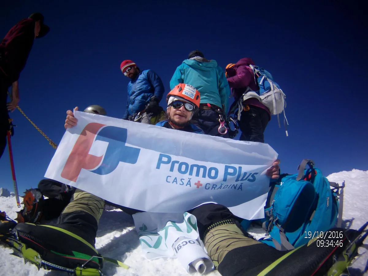 Trei alpiniști ținând un steag Promo Plus pe un vârf de munte